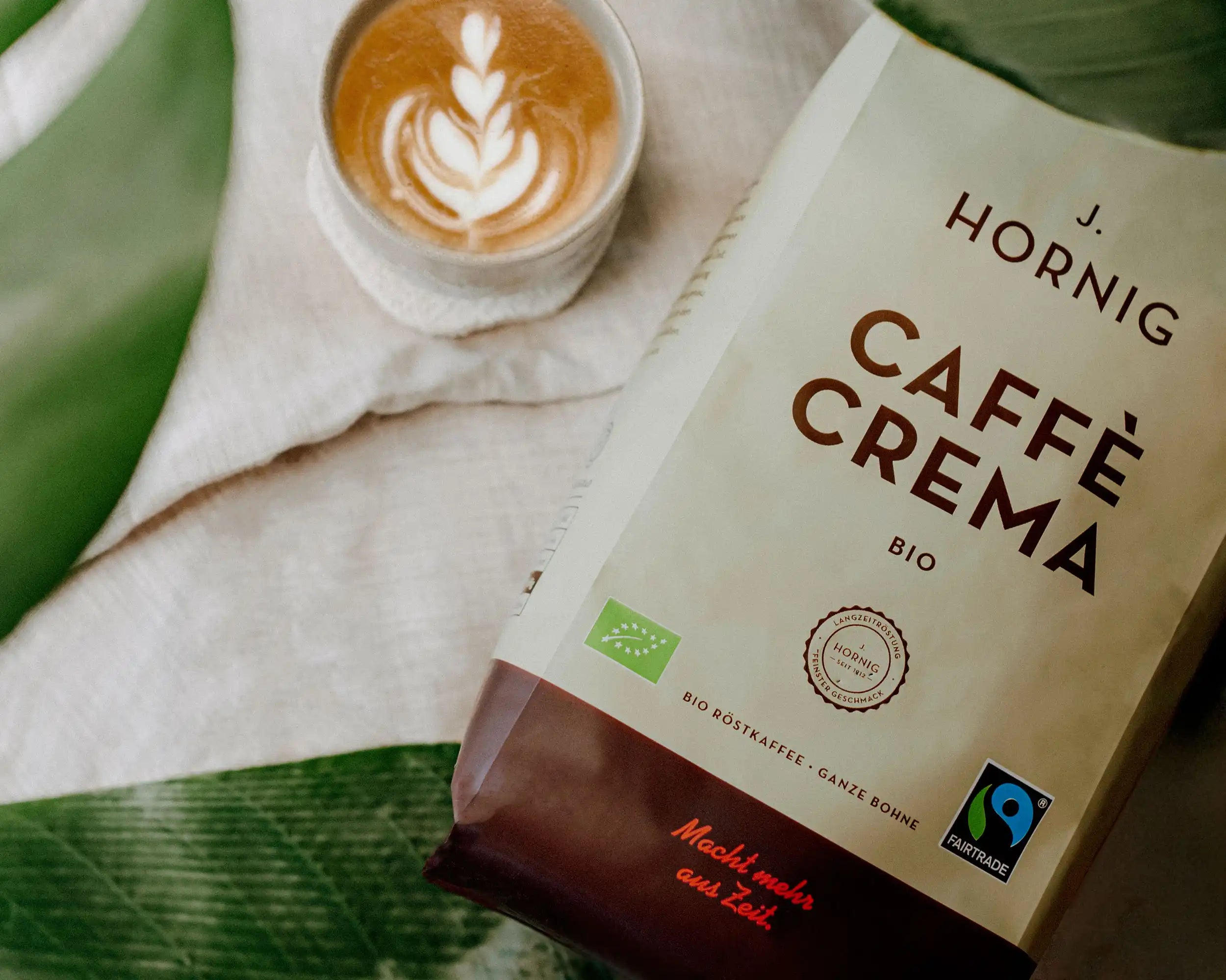 Eine Packung J. Hornig Caffe Crema Bio liegt auf einem Tisch mit einem Cappuccino.