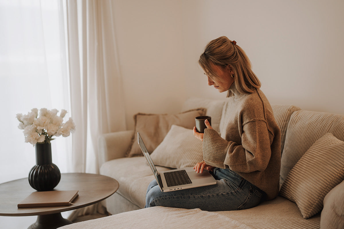 Eine Frau sitzt mit einer Tasse Kaffee und einem Laptop auf einem Sofa.